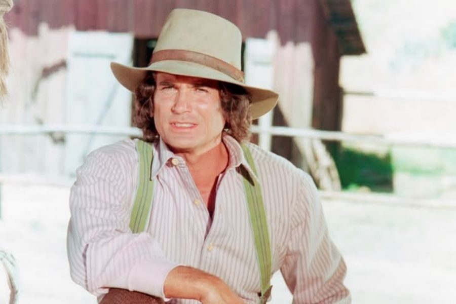 Man in a striped shirt and suspenders, wearing a wide-brimmed hat. He stands outdoors by a wooden structure, conveying a rustic, calm atmosphere.