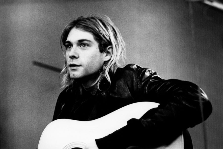 Black and white photo of a person with long hair playing an acoustic guitar. They wear a leather jacket and appear thoughtful, looking to the side.