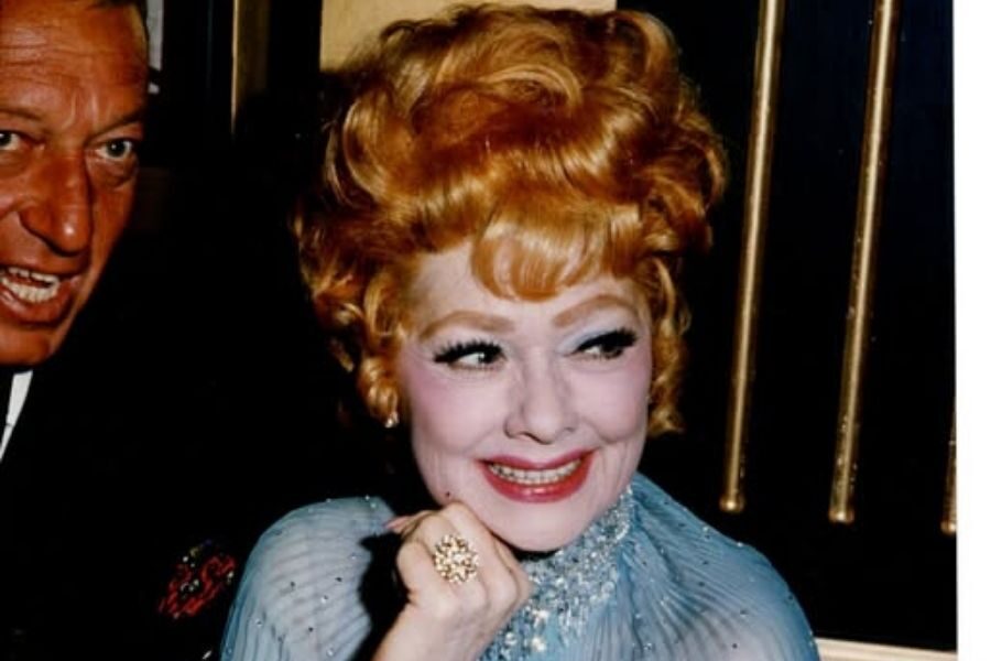 An elegantly dressed woman with red hair and a sparkling ring smiles warmly, conveying joy. A man in a suit is partially visible beside her.