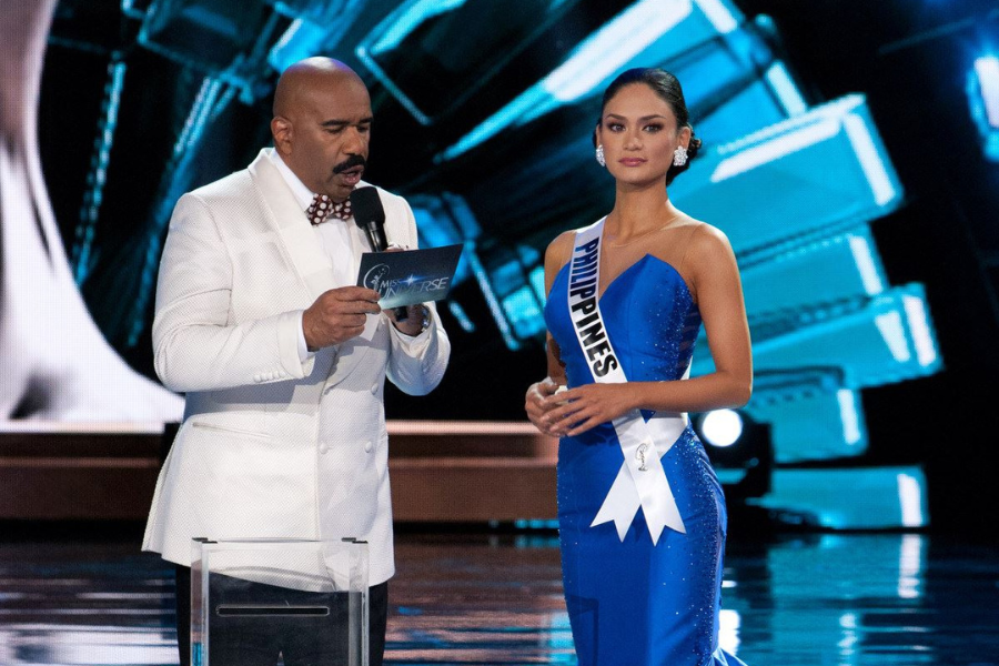 Host in a white suit, holding a microphone and card, stands next to a contestant in a blue gown and sash that reads "Philippines." The background is illuminated with blue and white lights, creating a formal and anticipatory atmosphere.
