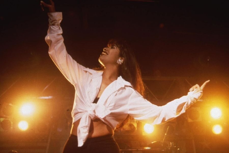 A woman in a white tied shirt joyfully performs on stage under warm, yellow stage lights. Her expression is vibrant and energetic.