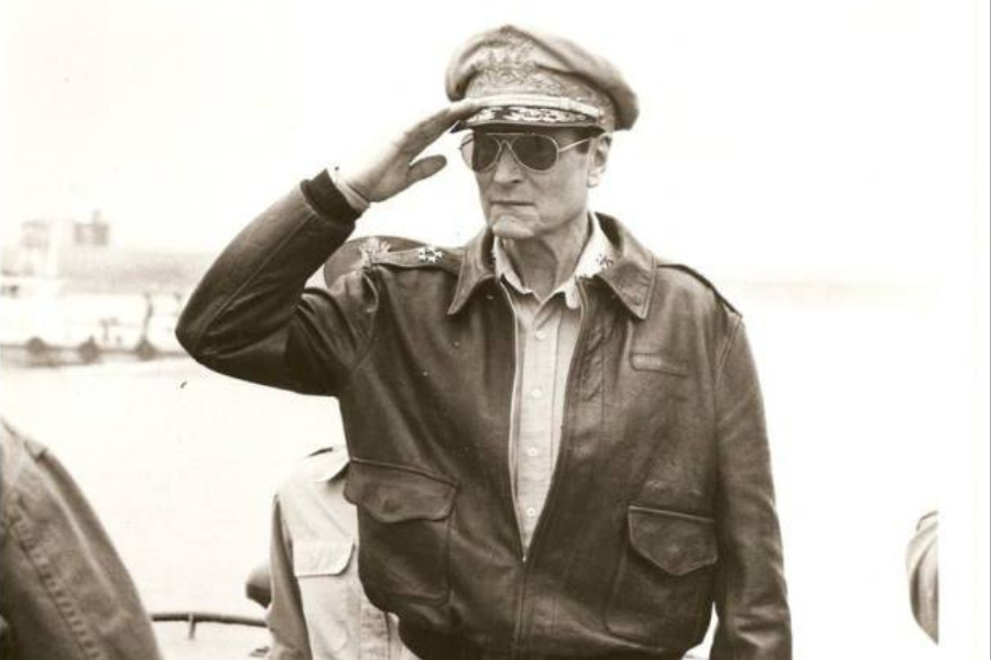 An officer in a leather jacket and cap salutes, exuding authority. He wears sunglasses and stands on a ship, with a calm sea as the backdrop.