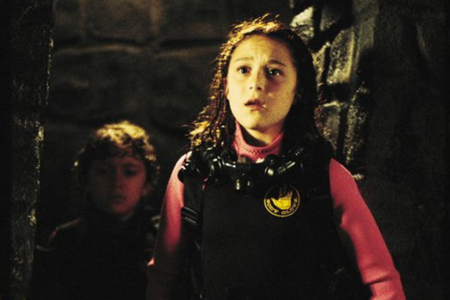A young girl with curly hair, wearing a pink and black wetsuit, stands in a dimly lit stone corridor, looking surprised. A child is blurred in the background.
