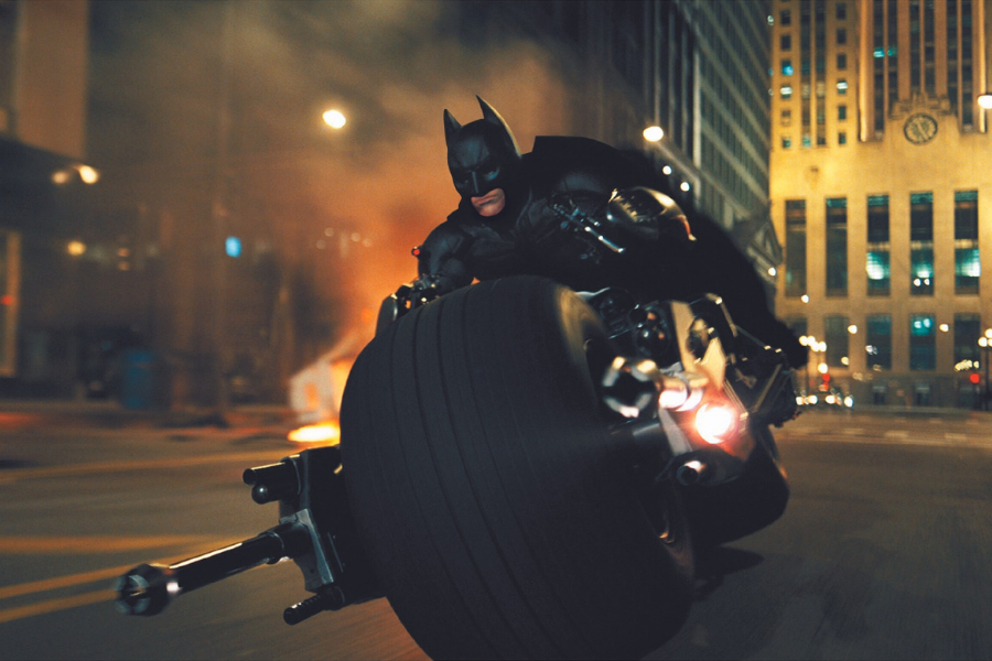 Batman rides a sleek, futuristic motorcycle at high speed through a dimly lit city street. Smoke and glowing lights add intensity and action.