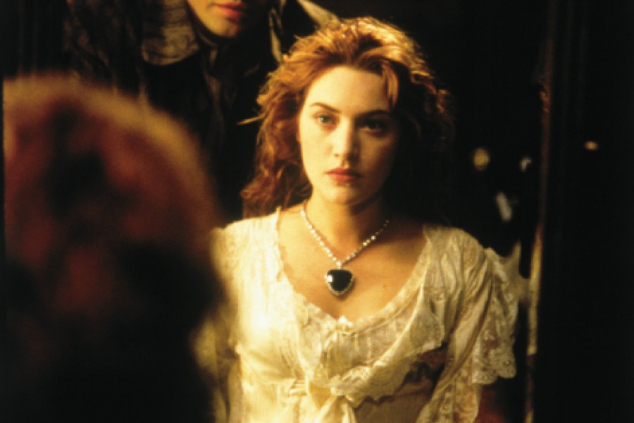 A woman with red hair, wearing a white dress and heart-shaped necklace, stands pensively in front of a mirror with a blurred reflection visible.