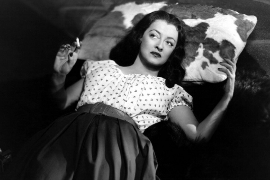 Black and white photo of a woman lying on a patterned cushion, holding a cigarette. She wears a printed blouse and a long skirt, gazing upwards thoughtfully.