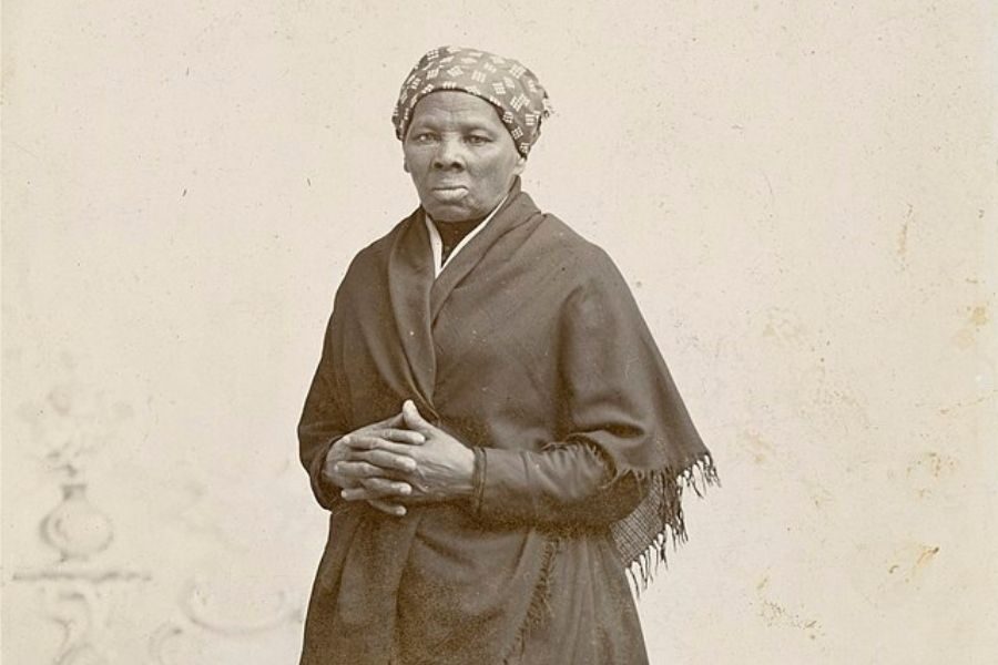 An older woman stands with her hands clasped, wearing a patterned headscarf and dark shawl. Her expression is calm and resolute, set against a plain background.