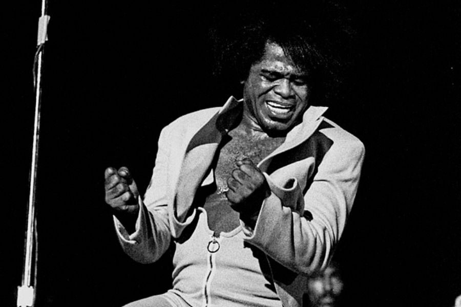 Energetic performer passionately singing on stage, wearing an expressive suit. His expressive face and clenched fists convey intense emotion. Black background.