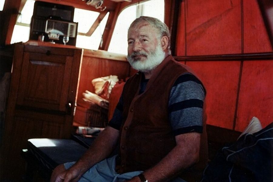 An older man with a white beard sits relaxed inside a boat cabin with warm wooden tones and soft sunlight. He wears a striped shirt and a vest.