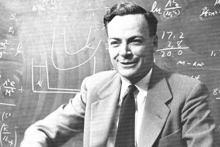 A person in a suit and tie smiles while standing in front of a blackboard filled with mathematical equations and graphs, suggesting a scholarly atmosphere.