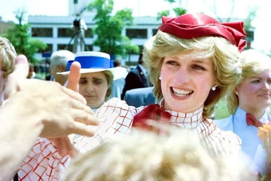 Smiling woman with short blonde hair, wearing a red hat and checkered outfit, greets a crowd. Others in the background look on. Positive atmosphere.