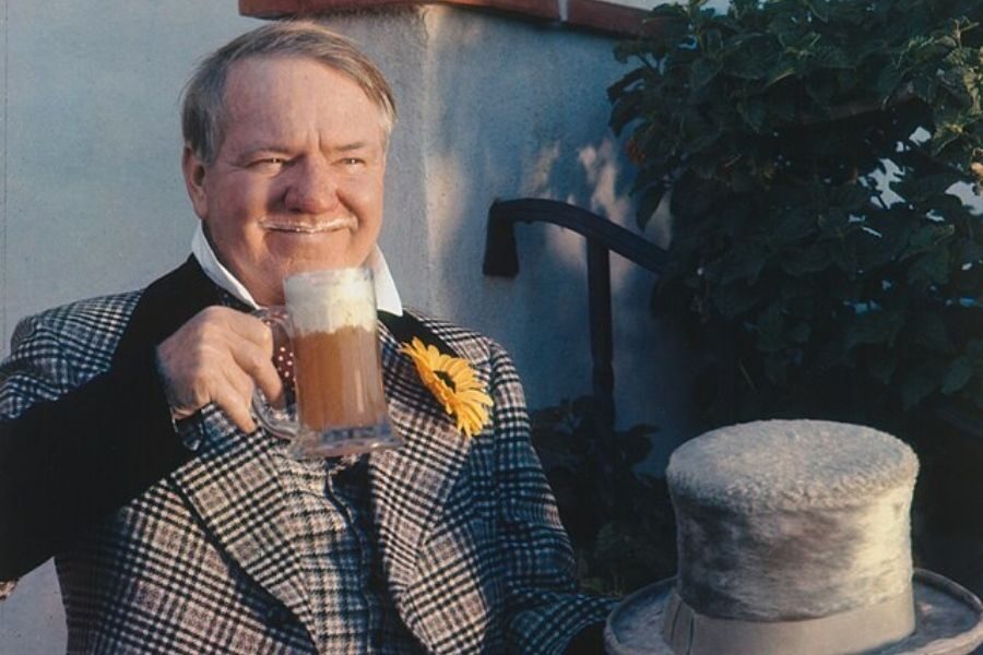 Older man in a plaid suit with a yellow flower, holding a frothy beer mug and a felt hat. He smiles warmly, exuding a relaxed, cheerful vibe.