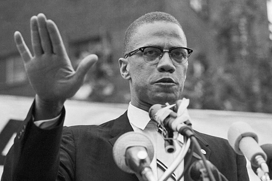 A man in a suit and glasses stands behind multiple microphones, raising his hand in a gesture. The scene is serious, conveying a sense of determination.