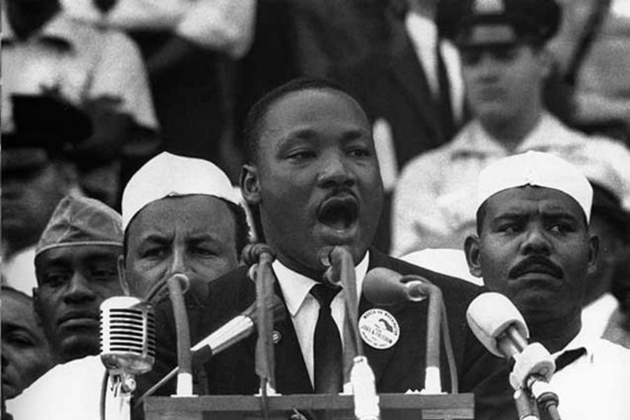A man passionately speaks at a podium surrounded by microphones, with a crowd in the background. His expression is focused and determined.