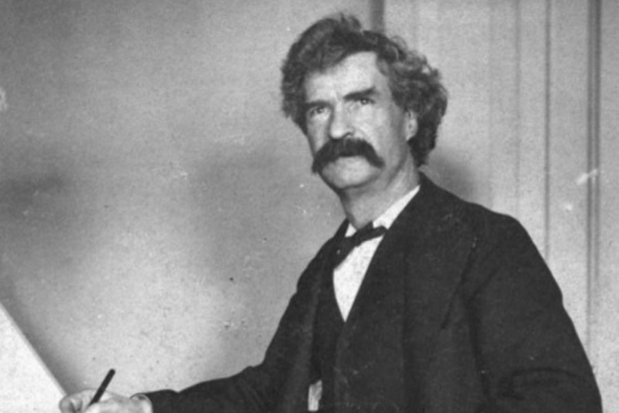A man with curly hair and a thick mustache, dressed in a dark suit, sits at a table writing with a pen, conveying a serious and focused tone.