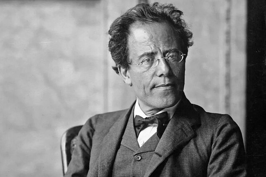 A man with glasses and tousled hair sits in a formal suit. He gazes slightly to the right, conveying thoughtfulness. The background is a plain wall.