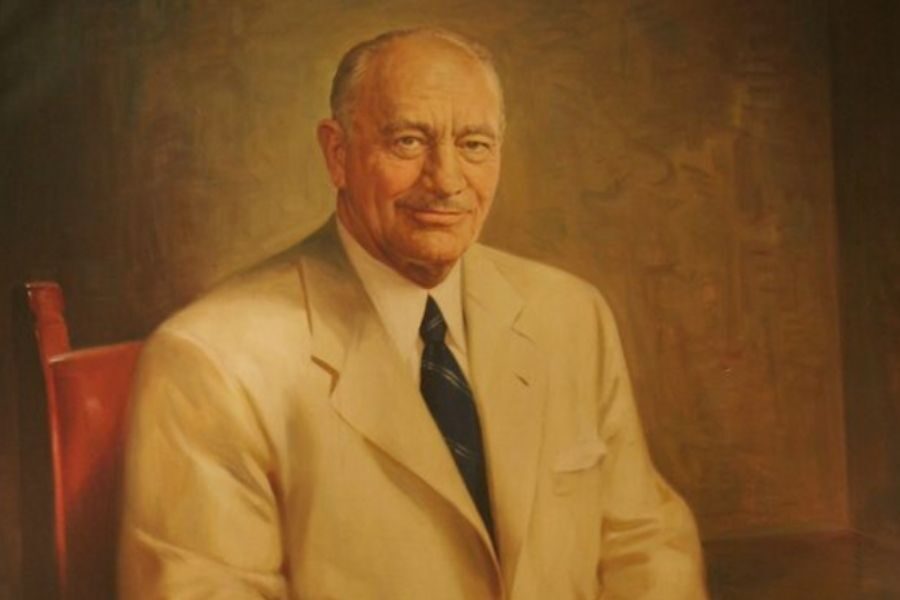 Portrait of an older man in a beige suit with a blue tie, seated on a red chair. He has a calm expression and the background is a warm brown.