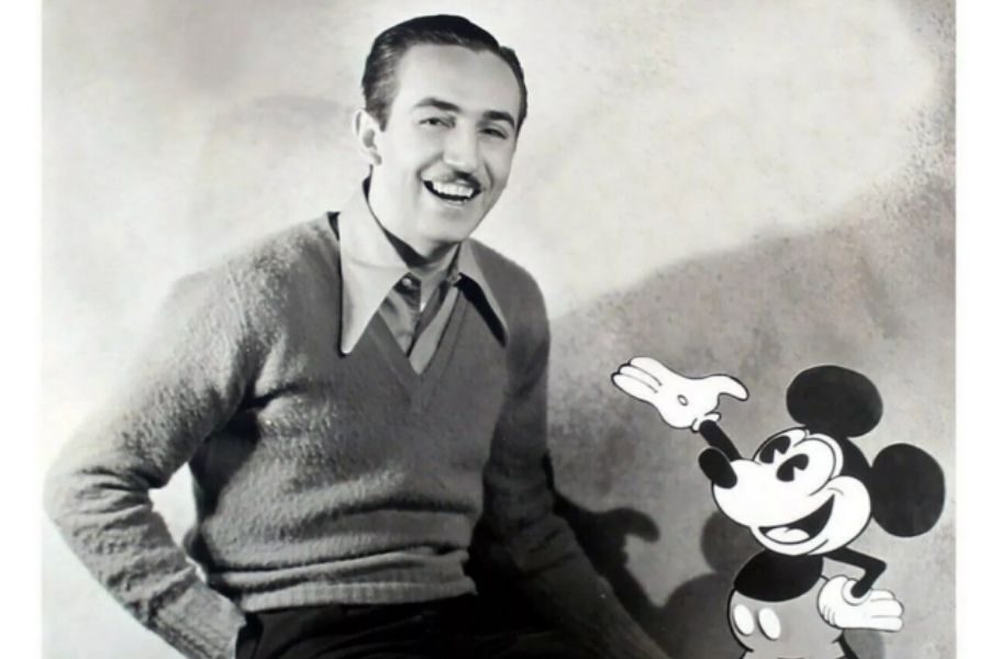 A smiling man with a mustache sits next to a cartoon character, Mickey Mouse, in a black-and-white photo. The image conveys a nostalgic and playful tone.