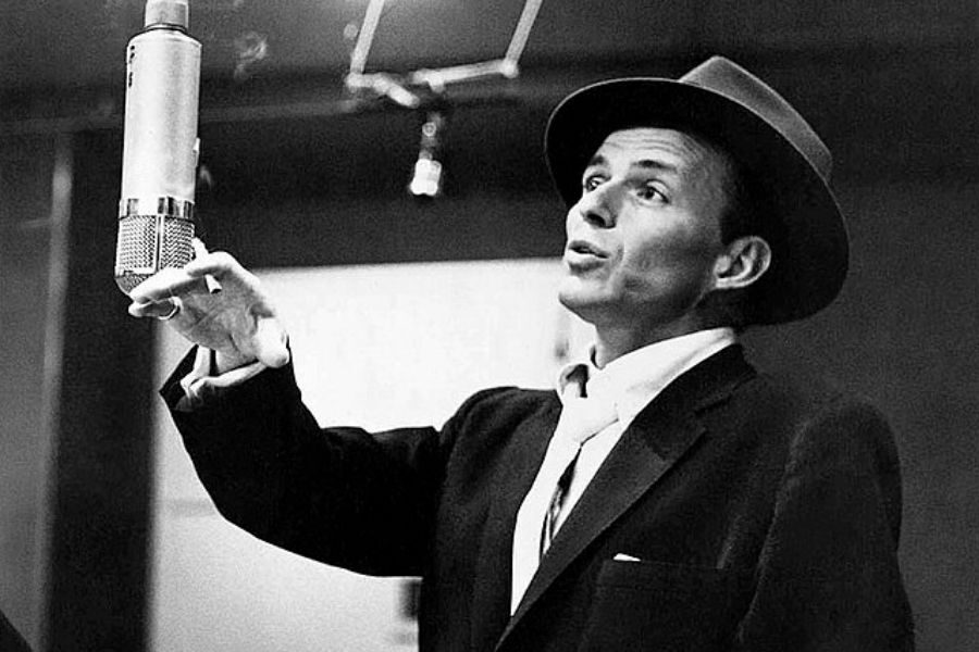 Black and white photo of a man in a suit and fedora, singing passionately into a vintage microphone in a recording studio. He appears focused and expressive.