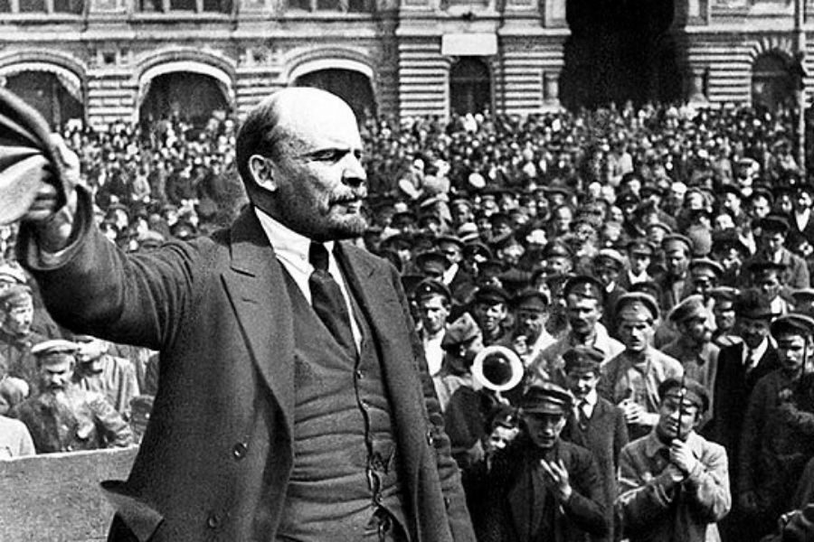 A man passionately addresses a large crowd outdoors. He holds a hat in one hand, projecting authority. Behind him, many people watch intently.