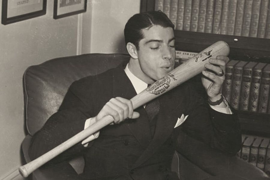 A man in a suit sits in an armchair, holding and kissing a signed baseball bat in a library. The setting is nostalgic, conveying admiration and passion.