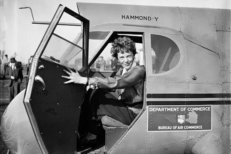 A woman with curly hair sits inside an open aircraft door, smiling at the camera. The plane has a "Department of Commerce" sign, exuding a sense of accomplishment.