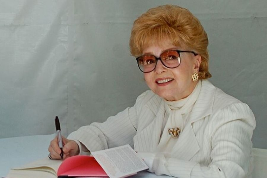 An older woman with short, reddish hair and glasses smiles while signing a book. She wears a white suit, exuding a friendly, welcoming demeanor.