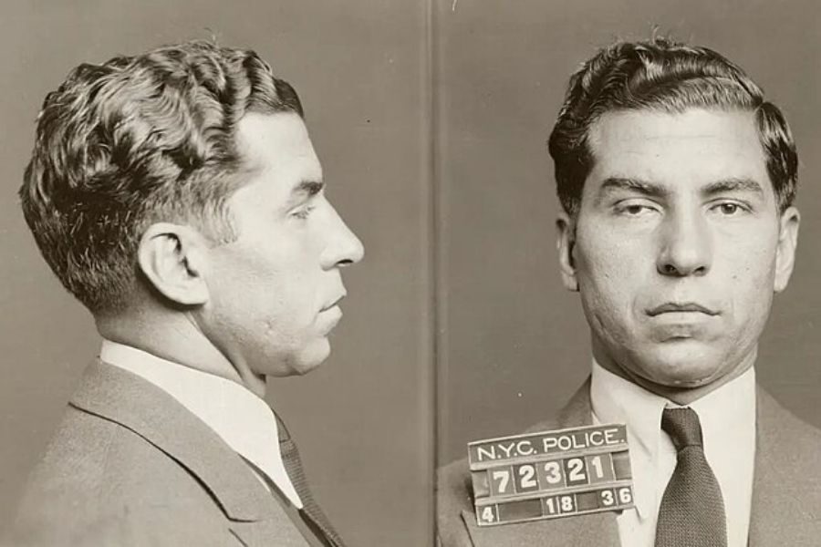 A sepia-toned mugshot shows a man in a suit facing left and looking directly at the camera. His expression is serious. The background is plain.