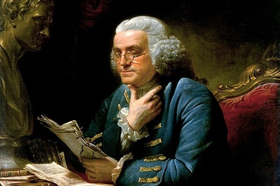 A distinguished older man with white hair and glasses reads papers, dressed in a blue coat. A bust and books are nearby, creating a scholarly atmosphere.