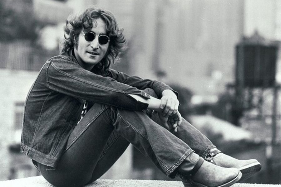 A person wearing a denim jacket, jeans, and sunglasses sits casually on a rooftop. They look relaxed, with a slight smile, against a blurred cityscape.
