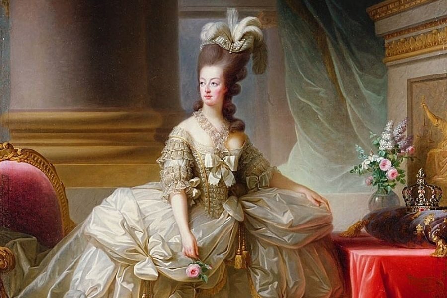 A regal woman in an ornate gown and elaborate hairpiece sits beside a table with flowers and a crown on red fabric. The setting is opulent and elegant.