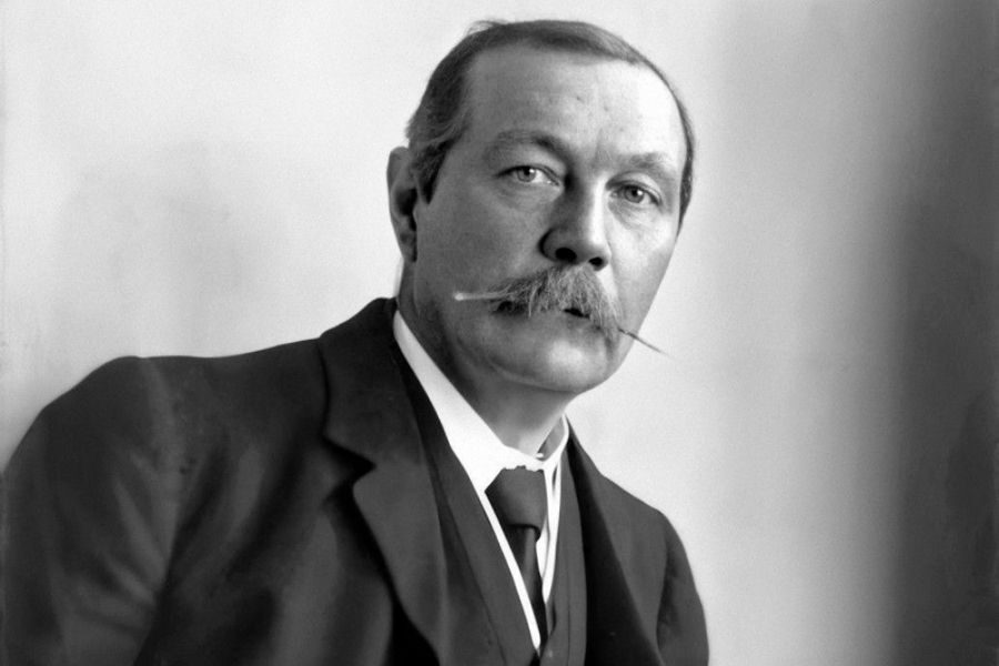 An older man with a prominent mustache wearing a dark suit and tie, gazes thoughtfully. The background is plain, highlighting his focused expression.