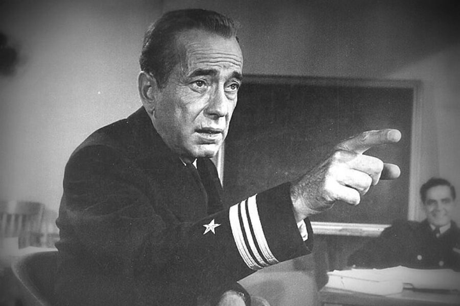 Grayscale image of an intense, uniformed man pointing, evoking drama. A blurred figure sits in the background near a chalkboard, creating tension.