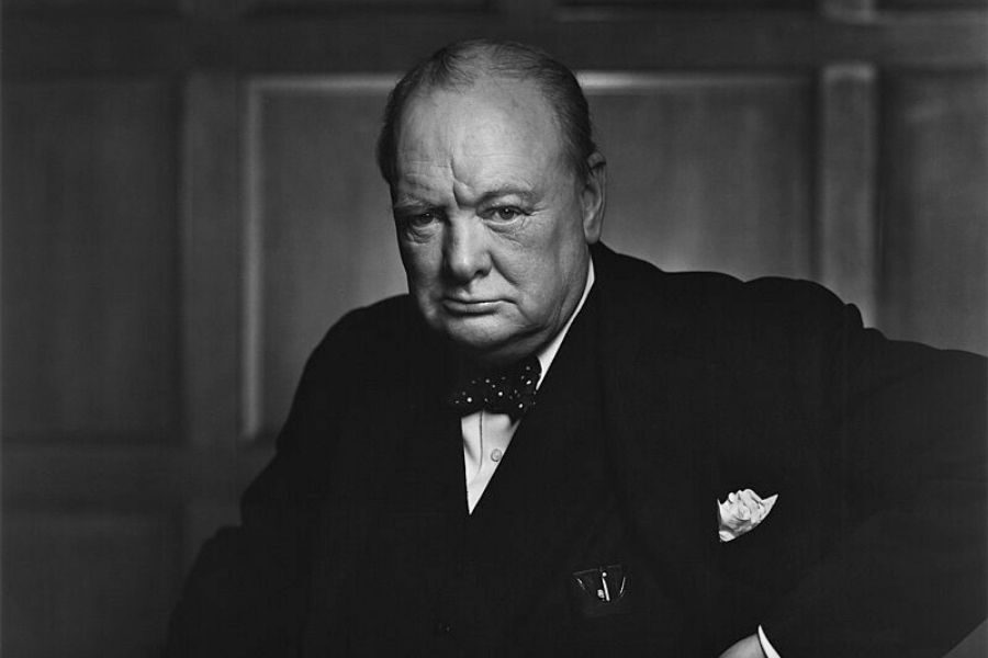 Black and white portrait of a serious, older man in a suit and bow tie, with a determined expression, standing against a wooden panel background.