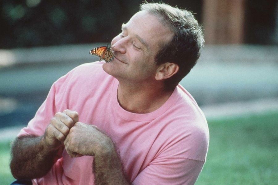 A person wearing a pink shirt smiles gently with closed eyes as a butterfly rests on their nose, creating a peaceful and joyful atmosphere in a garden setting.