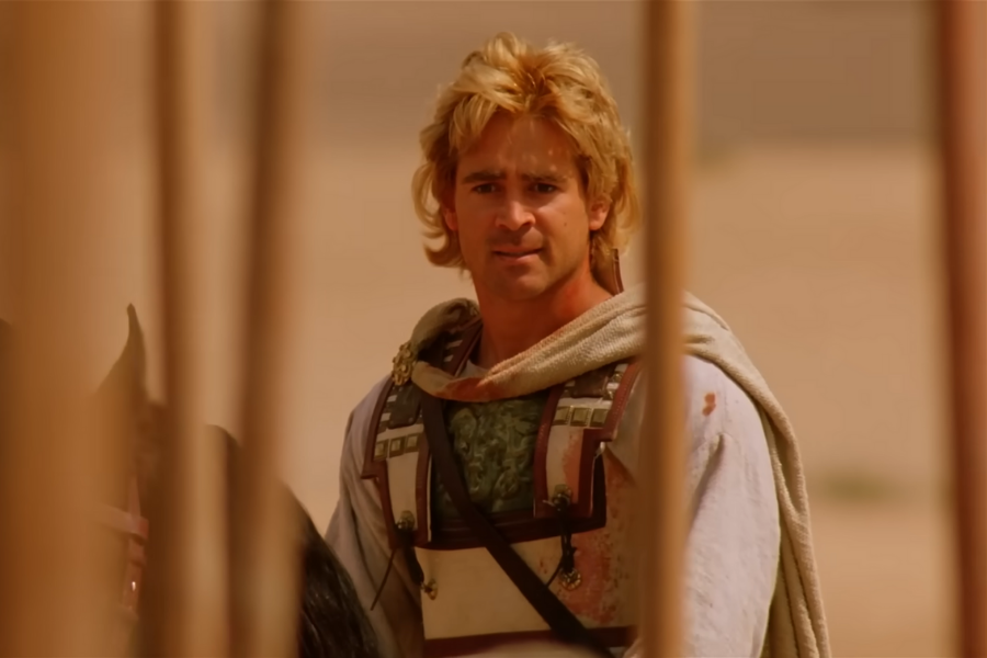 A man with blond hair and a concerned expression stands in a desert, wearing ancient armor and a cloak. Wooden poles frame him in the foreground.