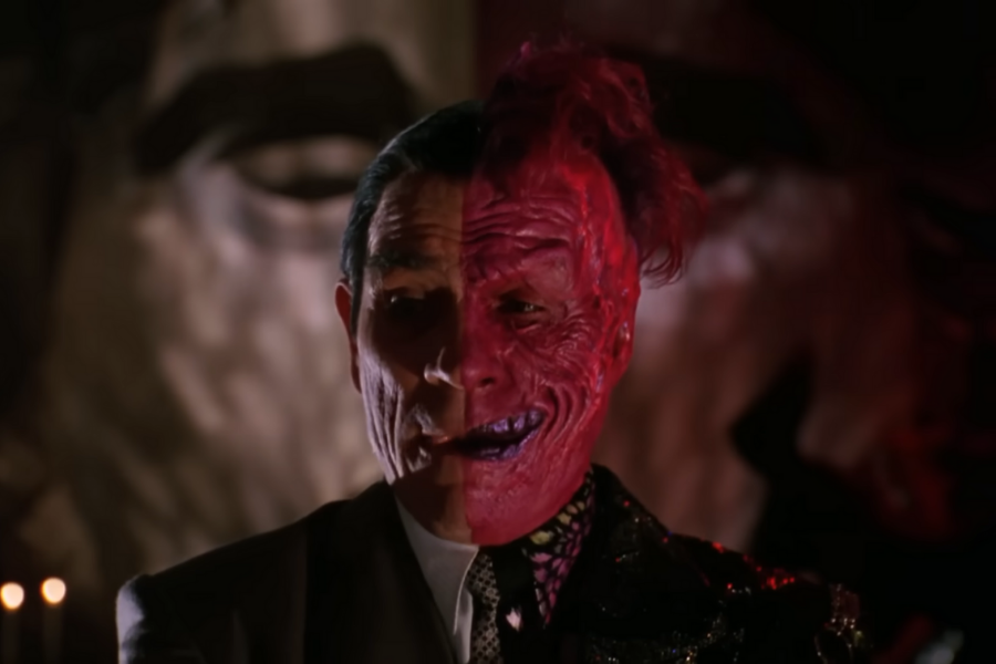 A man with a dual-toned face, one side normal and the other scarred and red, wearing a suit, stands against a dimly lit, dramatic background.