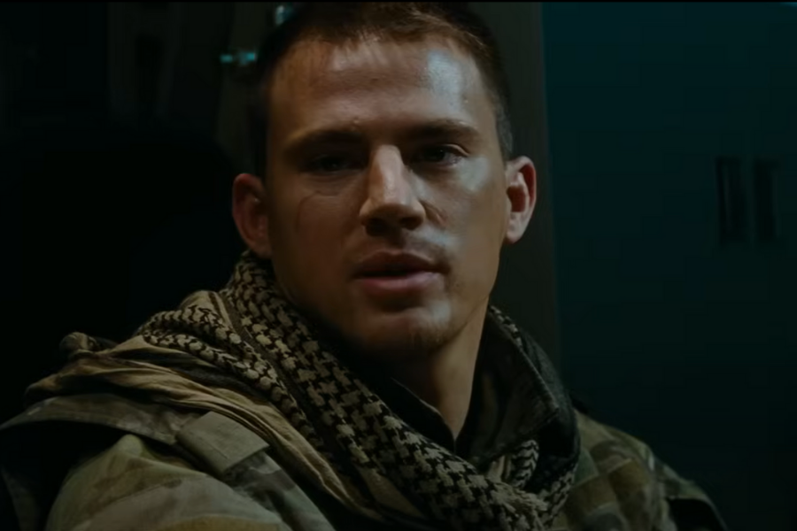 A man with short hair and a serious expression, wearing a camouflage uniform and a patterned scarf, is seated in dim lighting, conveying a tense, introspective mood.