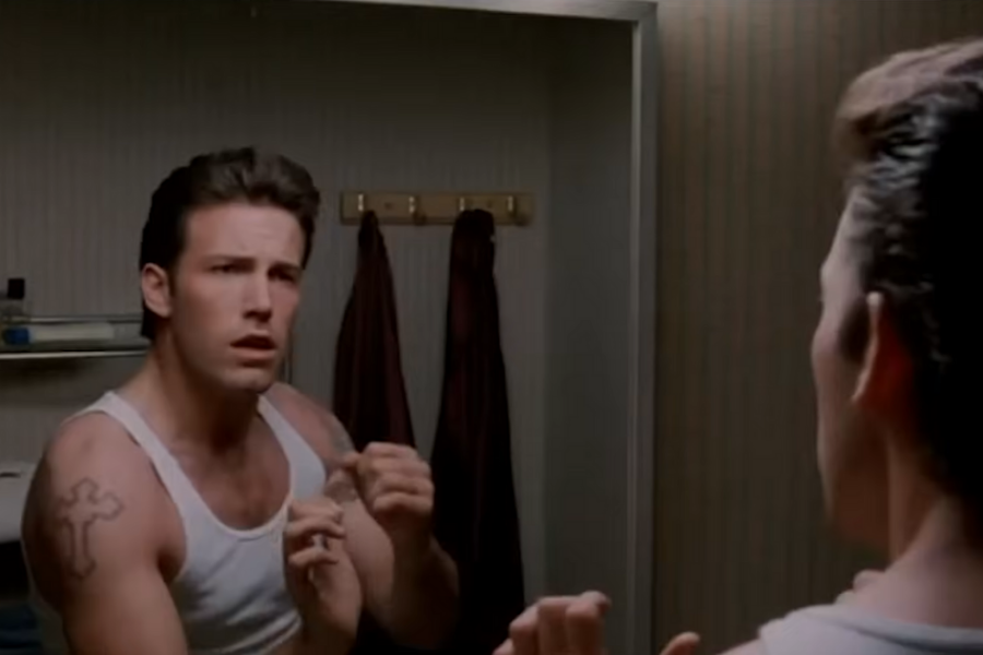 Man in a white tank top with a tattoo looks worried while facing a bathroom mirror. Towels hang on hooks behind him.