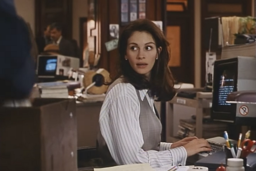 A woman with long brown hair looks surprised while seated at a cluttered office desk, typing on a computer. The background shows a busy office environment.