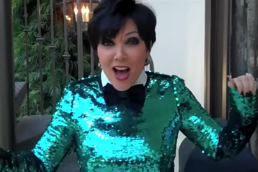 A person excitedly poses indoors, wearing a vibrant, sparkly green top and a black bowtie. The background features greenery and warm lighting.