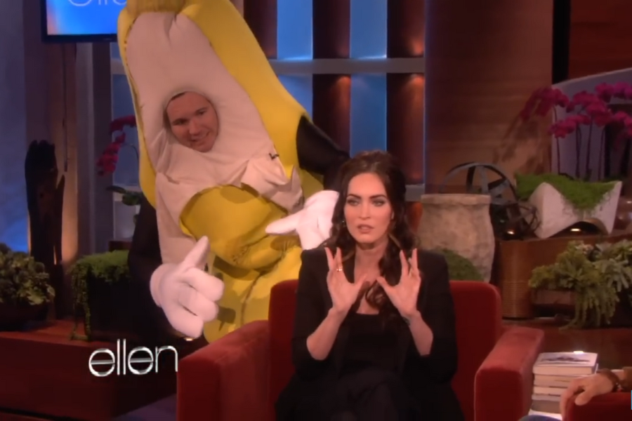A woman, appearing surprised, speaks while sitting on a talk show set. Behind her, a person in a banana costume is ready to surprise her. The atmosphere is fun and lively.
