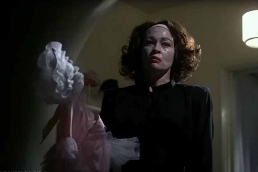 A woman with a stern expression stands in dim lighting, wearing a face mask and headband. She holds a doll in a pink dress, creating an eerie mood.