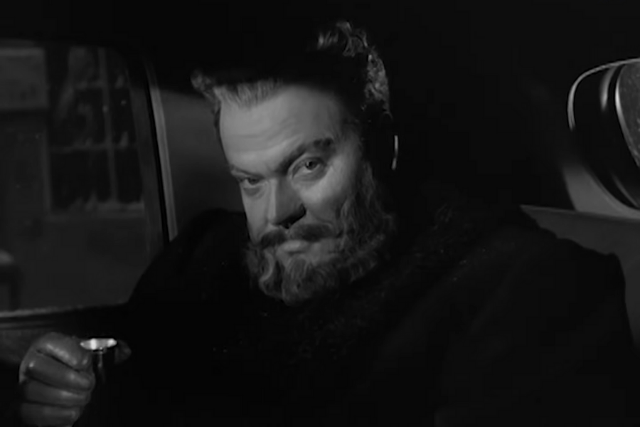 Bearded man in a dark coat sits in a dimly lit car, gazing intently with a slight smile, holding a small cup, creating a mysterious atmosphere.