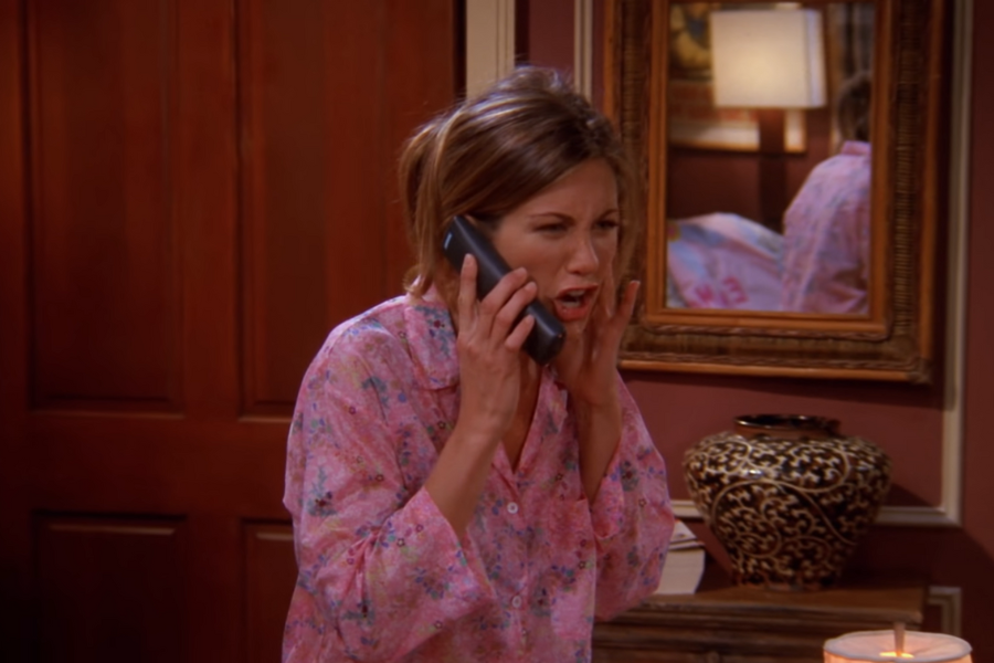 Woman in pink floral pajamas is on the phone, looking surprised or frustrated. Background shows a wooden door, ornate mirror, and decorative vase.