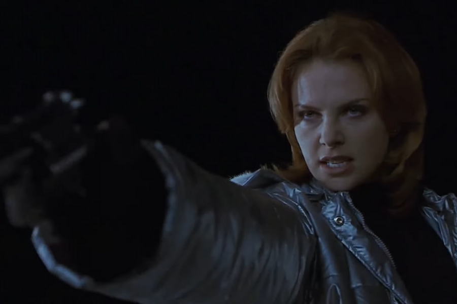 A woman with determined expression aiming a gun off-camera. She wears a shiny silver coat against a dark background, evoking suspense and tension.