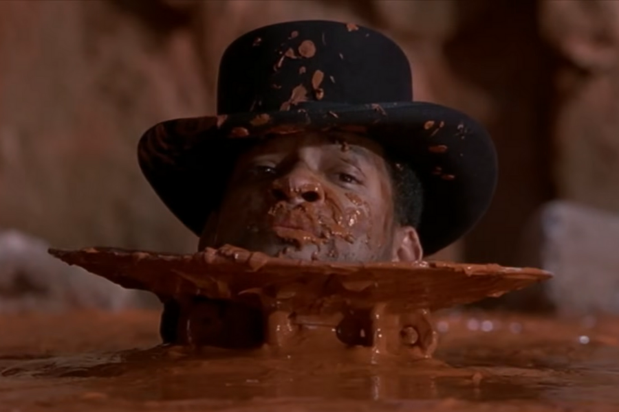 A person's face and top hat are splattered with mud, and they are partially submerged in a mud pool. The expression appears humorous amidst muddy surroundings.