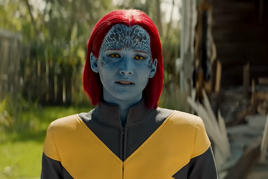 Blue-skinned character with red hair and intricate face patterns wearing a yellow and black suit stands outdoors, conveying a serious expression.