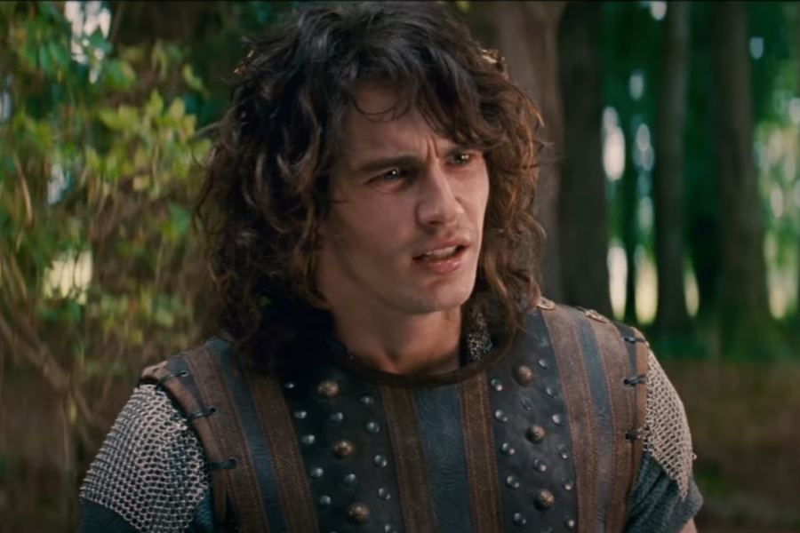 A person with curly hair, wearing medieval leather armor with studs, stands in a forest, looking confused. The light filters through the trees, adding a dramatic tone.