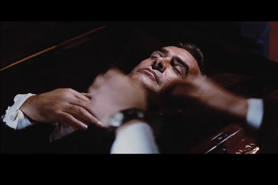 A man lies in a casket with eyes closed and hands clasped. The scene is dimly lit, conveying a solemn and tense mood, with blurred movement nearby.